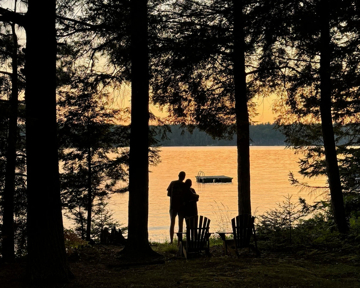 Two people standing together beside Adirondack chairs, overlooking a lake through the trees.
